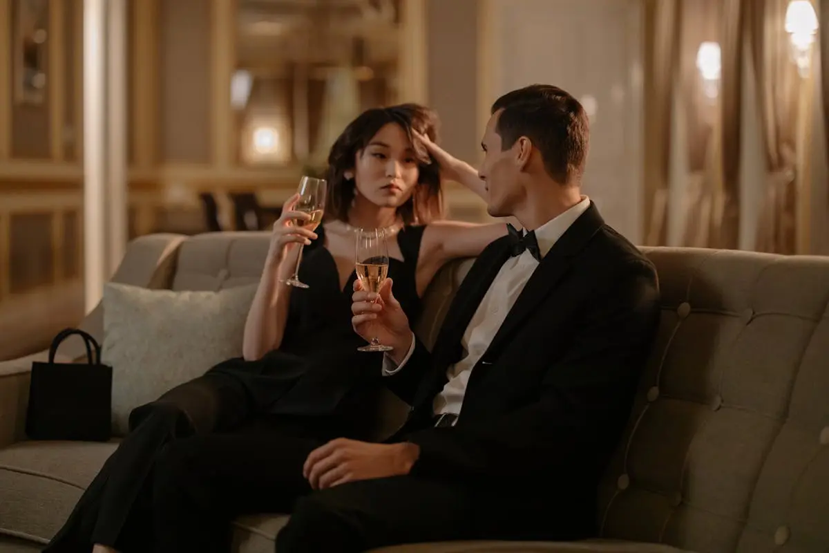 A stylish couple in formal wear enjoying champagne on a cozy sofa in an elegant indoor setting.