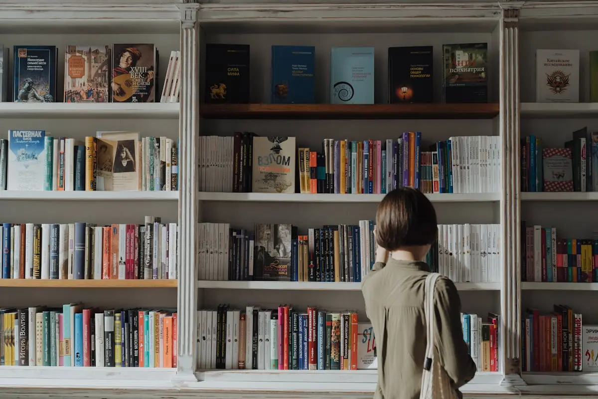 Woman in Beige Long Sleeve Shirt Standing in Front of White Wooden Book Shelf