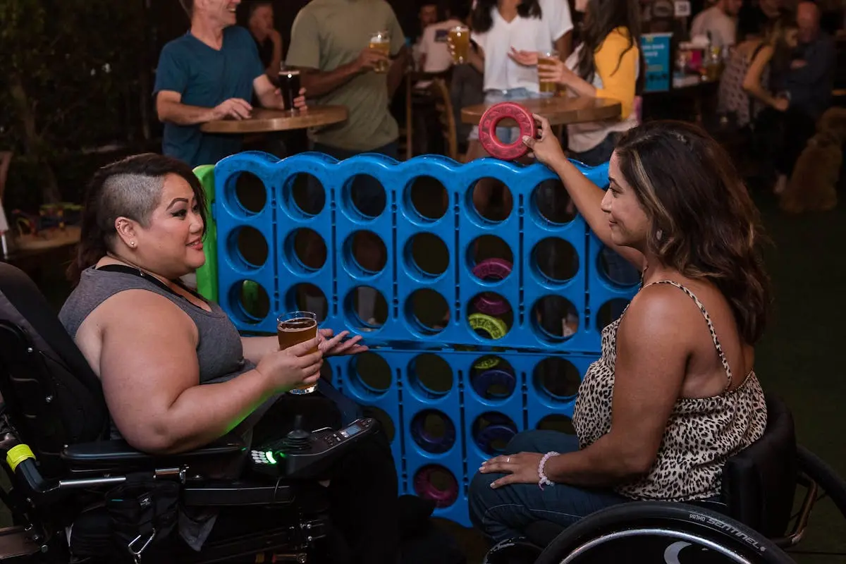 Two women in wheelchairs playing connect four at an indoor bar setting, enjoying a lively social atmosphere.