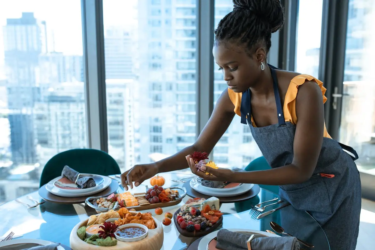 A woman arranges a charcuterie board with various cheeses and fruits on a glass table indoors.