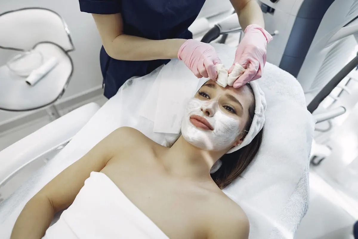 Unrecognizable crop female beautician in medical coat and latex gloves applying facial treatment of young woman resting on couch in modern spa studio