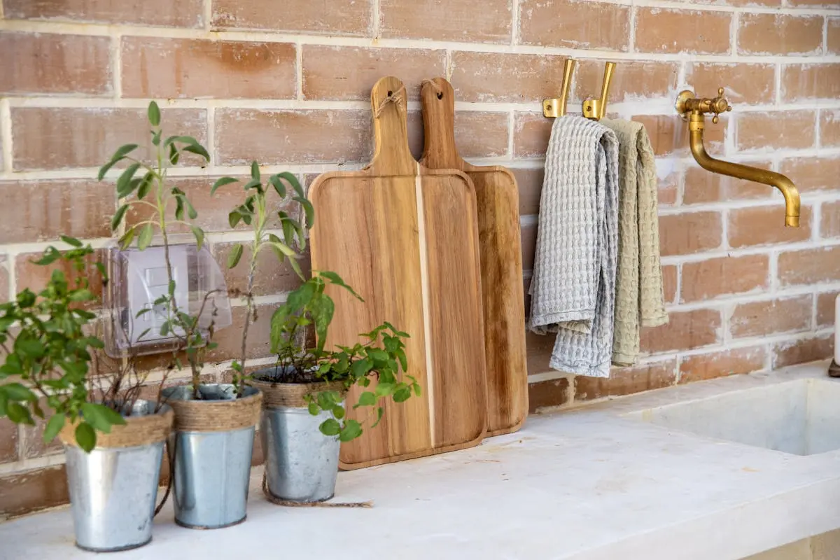 A rustic kitchen setup featuring herb plants, cutting boards, and a brass faucet against a brick wall.