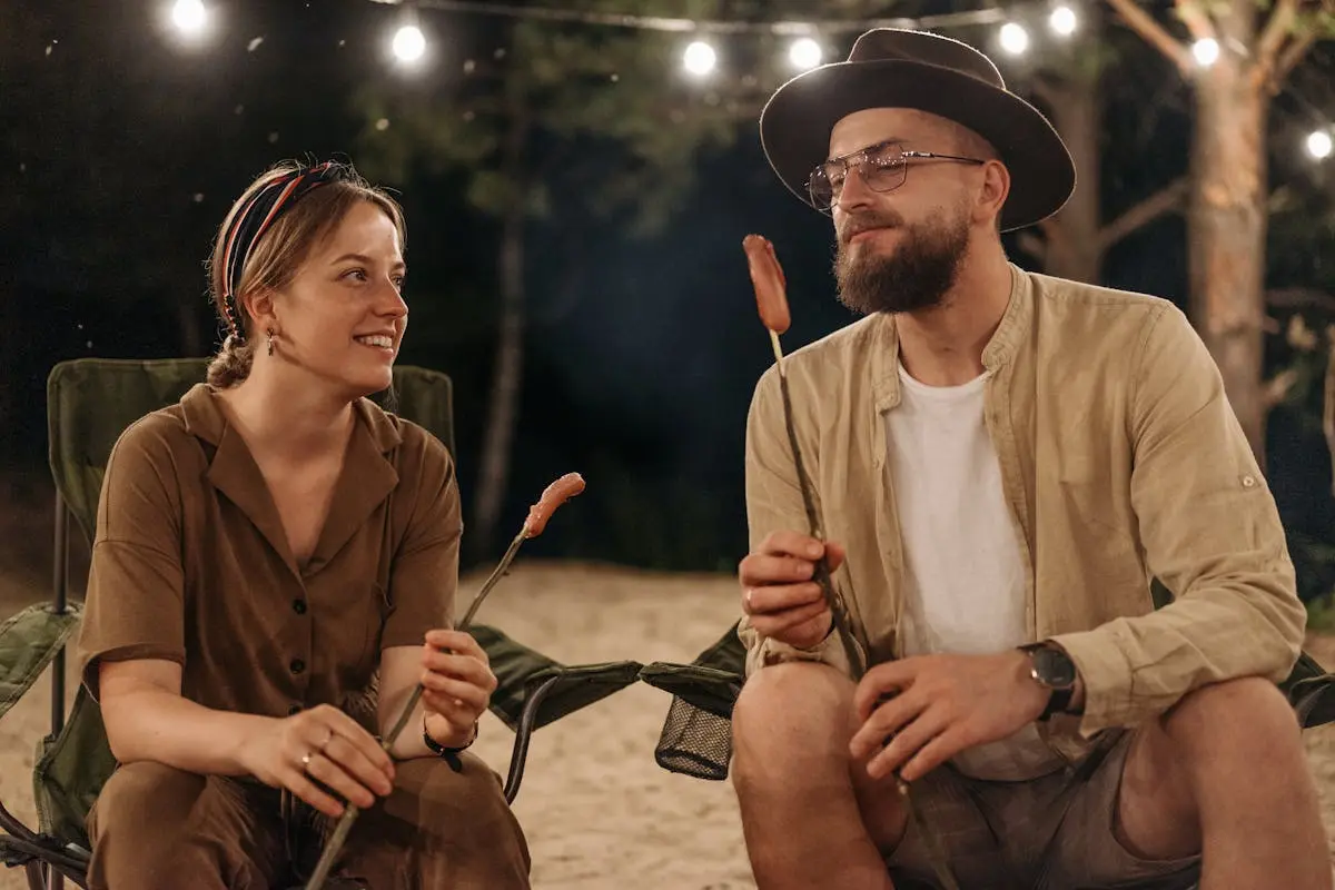 A couple enjoying a campfire at night, sitting in folding chairs and roasting food under string lights.