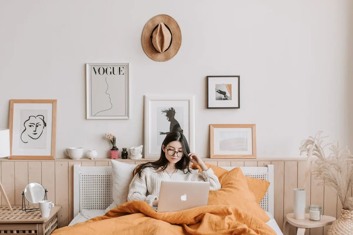 Young woman in cozy bedroom using a laptop, surrounded by stylish decor and art.