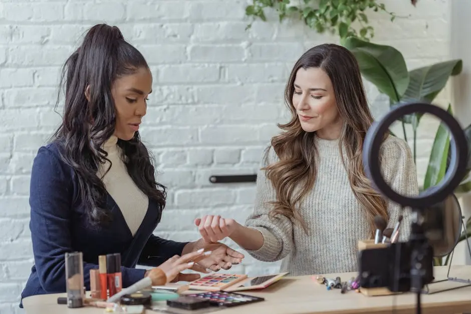 Young positive multiethnic female bloggers testing makeup products while sitting at table and recording vlog on smartphone placed on tripod with ring light