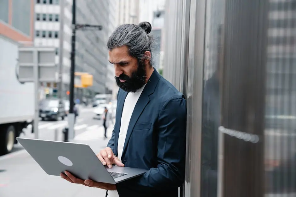 Bearded businessman focusing on laptop work in an urban outdoor setting.