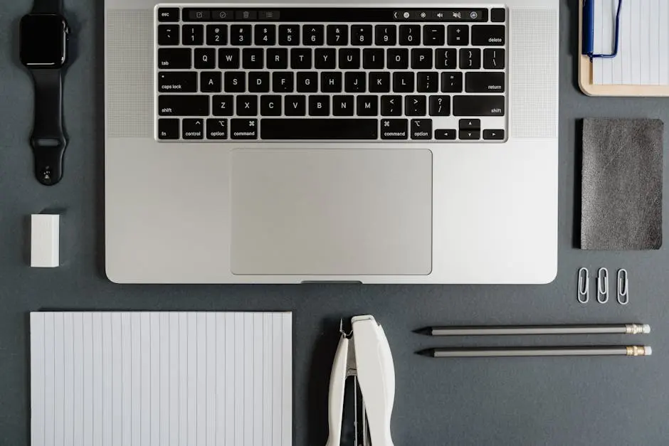 Top view of a sleek office desk with a laptop, notepad, smartwatch, and stationery essentials.