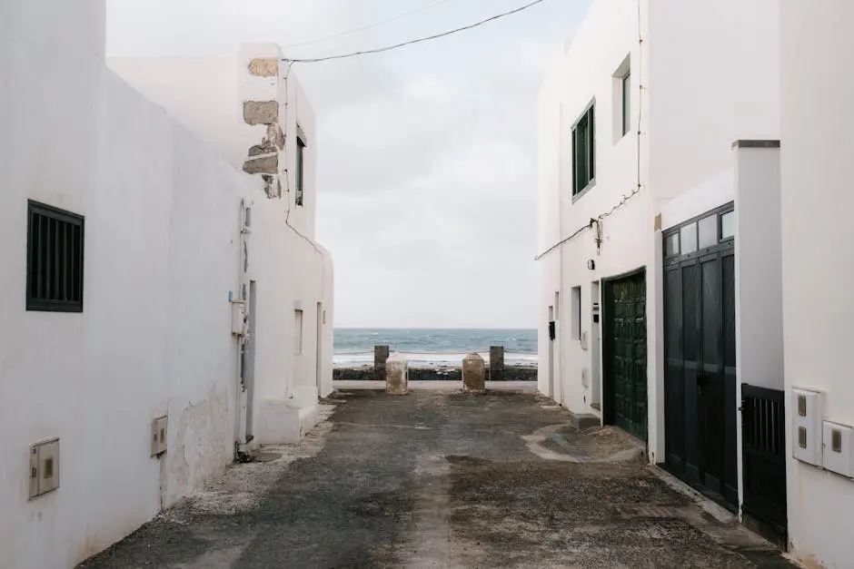 A serene coastal alleyway between white buildings leading to the ocean view on a cloudy day.