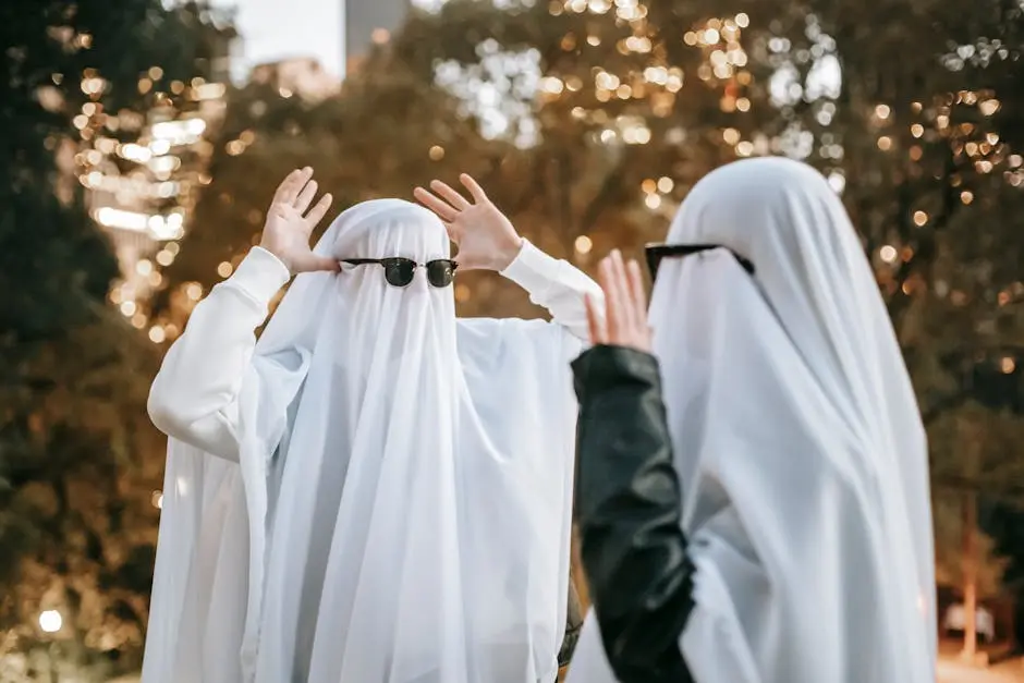 Cheerful anonymous couple with raised arms wearing ghost costumes and sunglasses looking at each other and having fun while standing in park with trees on blurred background during Halloween