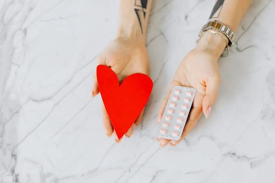 Close-up of hands holding a red heart shape and a blister pack of pills on a marble surface.