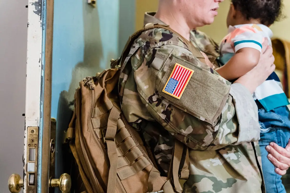 A soldier in camouflage uniform embraces a young child indoors, showing family warmth.