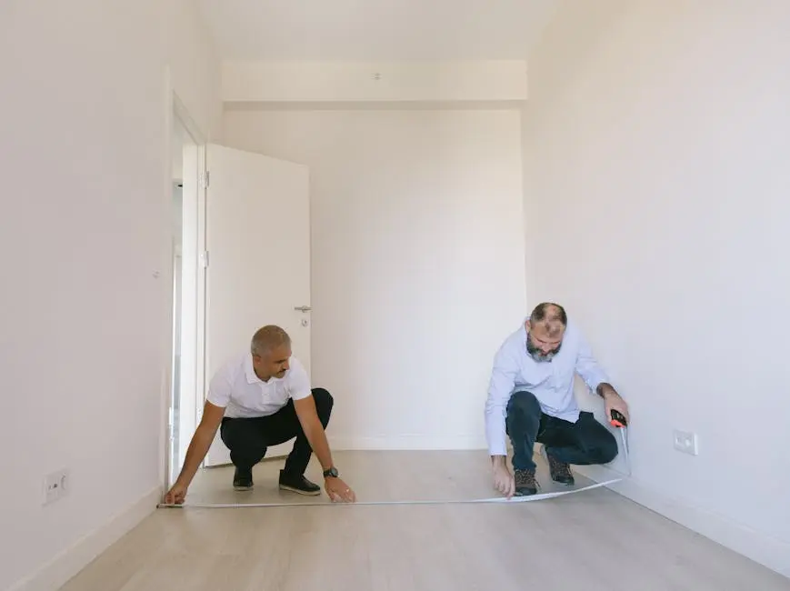 Two men measuring dimensions of a room with white walls and door, planning an interior renovation.