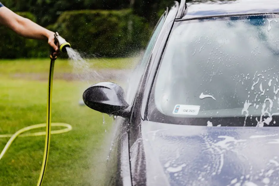 Person washing a car with a hose outdoors, creating suds on a sunny day.