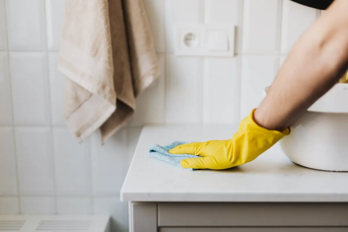 Close-up of a person wearing yellow gloves wiping a bathroom counter with a cloth.