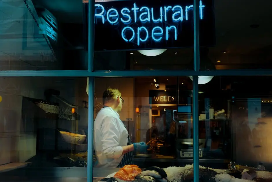 Chef in a restaurant behind a window with a neon 'Restaurant Open' sign, preparing seafood.