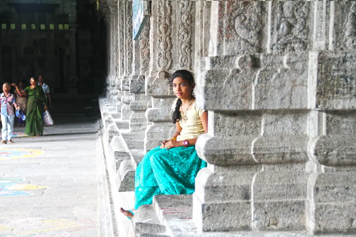 A teenage girl in colorful traditional attire sitting in a historic Kanchipuram temple, Tamil Nadu, India.