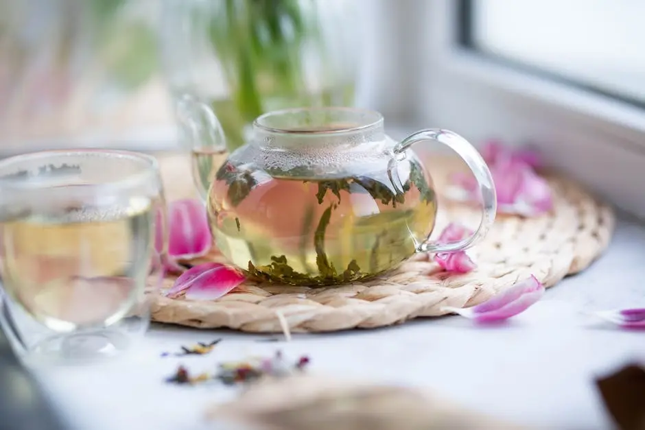 Transparent teapot with loose leaf tea placed on wicker mat with petals on white windowsill with glass on blurred background