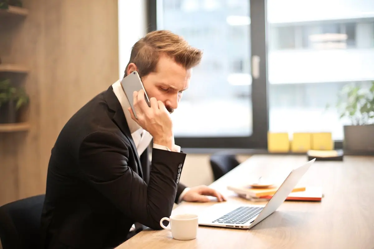 A businessman in a suit making a phone call while working on a laptop in a modern office setting.