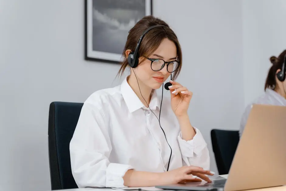 Young woman in glasses and headset providing customer support at a laptop in an office setting.