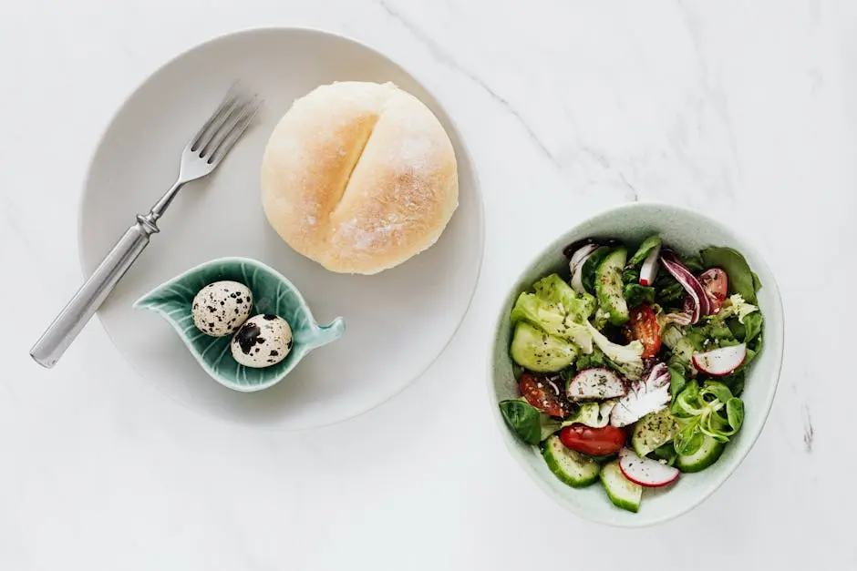 From above of healthy diet salad in bowl and plate with quail eggs fork and bun placed on white marble table