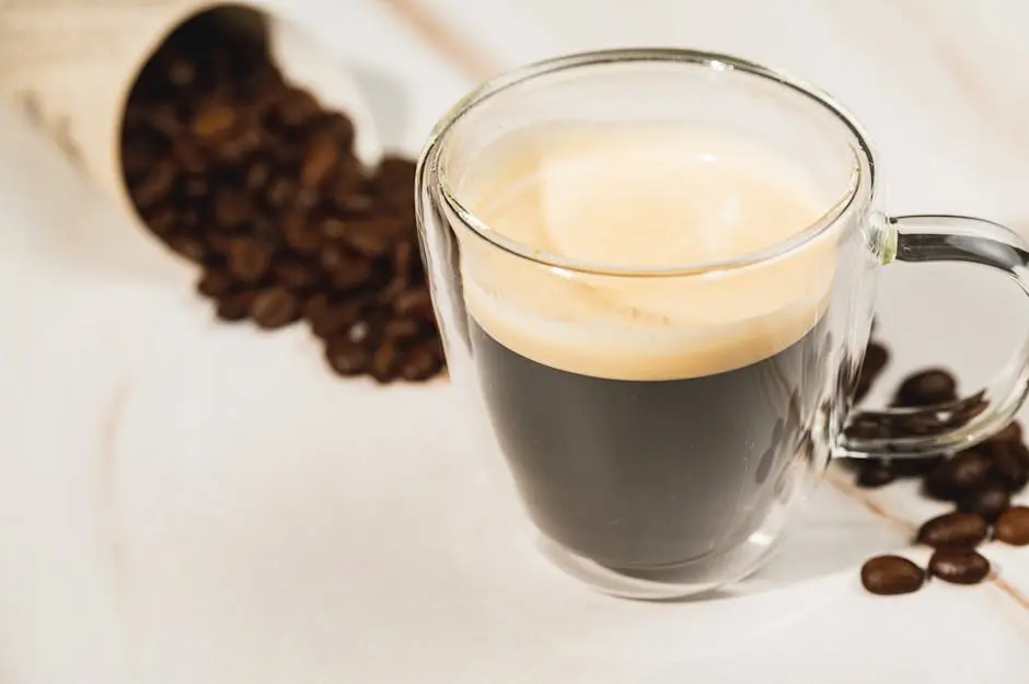 A clear glass cup of espresso on a white surface with scattered coffee beans.