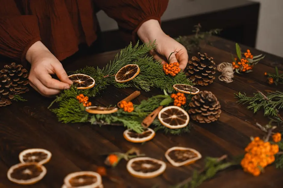 Hands arranging a seasonal wreath with dried oranges, pinecones, and foliage on a wooden table.