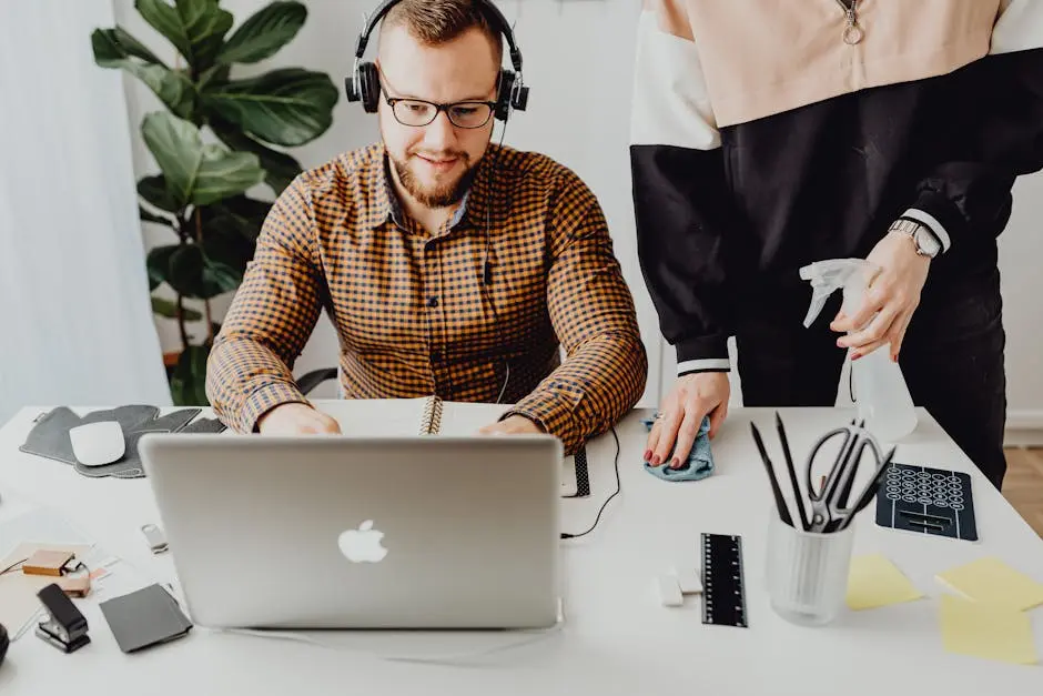 A man with headphones works on a laptop while another person cleans the desk in a modern office.